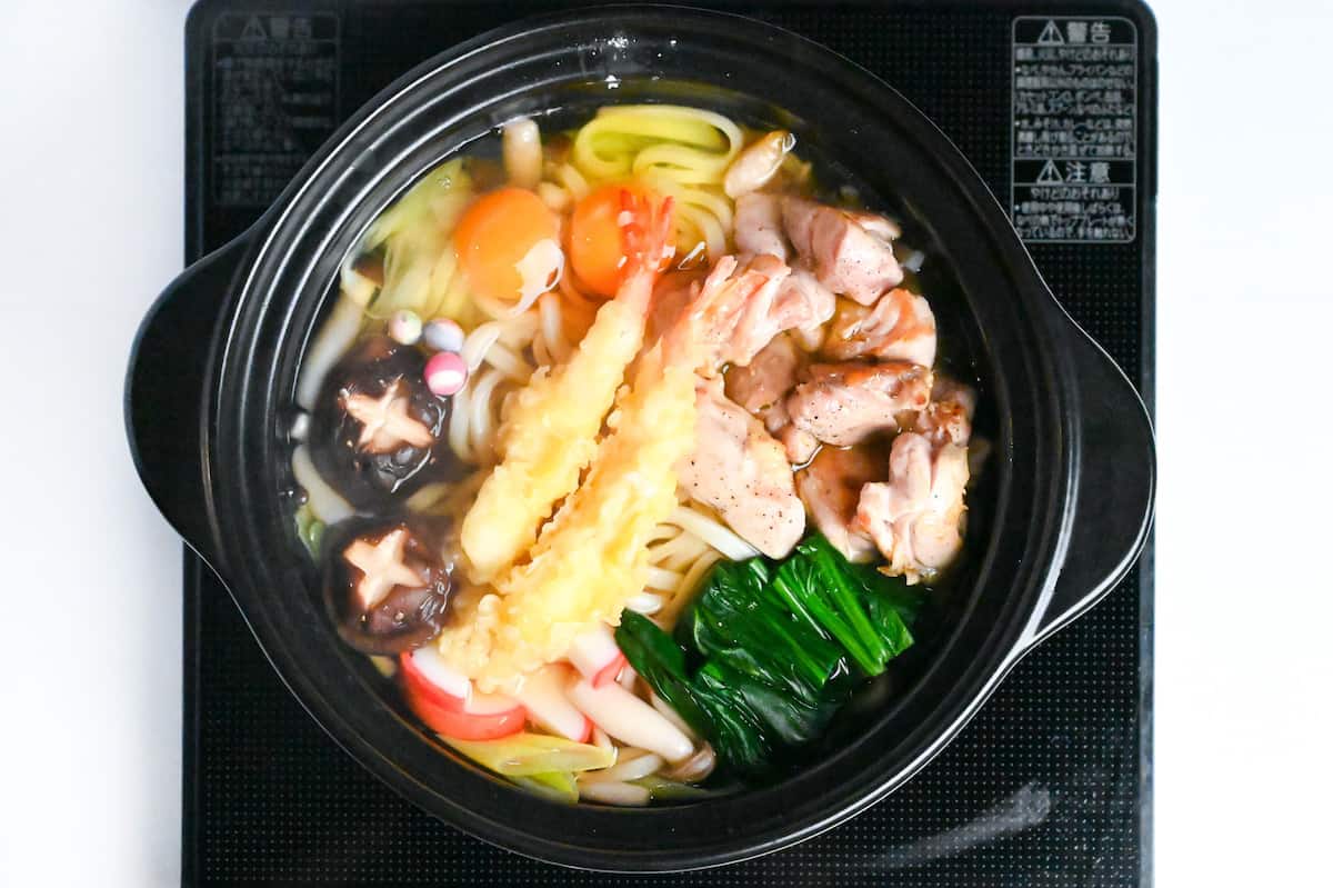 udon noodles, chicken, kamaboko (fishcake), fu, spinach and prawn tempura in dashi based soup in a black pot on the stove