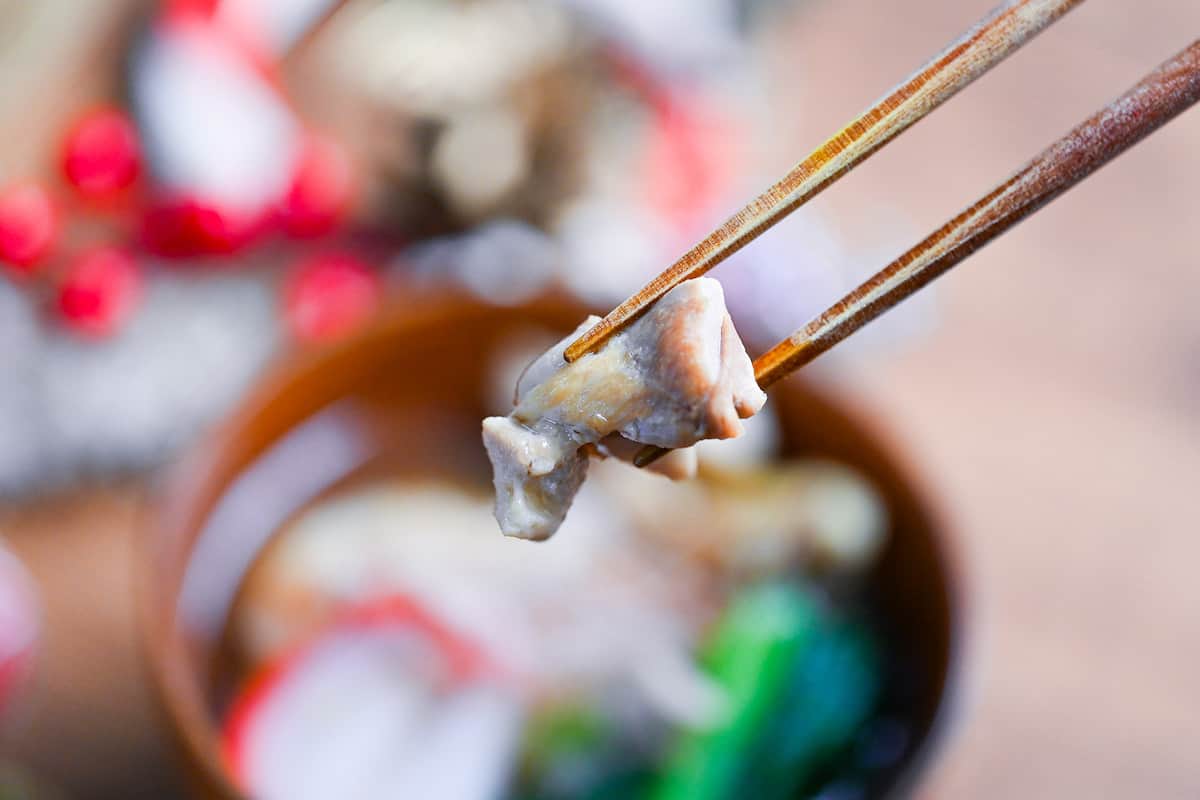 a piece of chicken held with wooden chopsticks from a bowl of Nagoya style ozoni (Japanese New Year's soup)