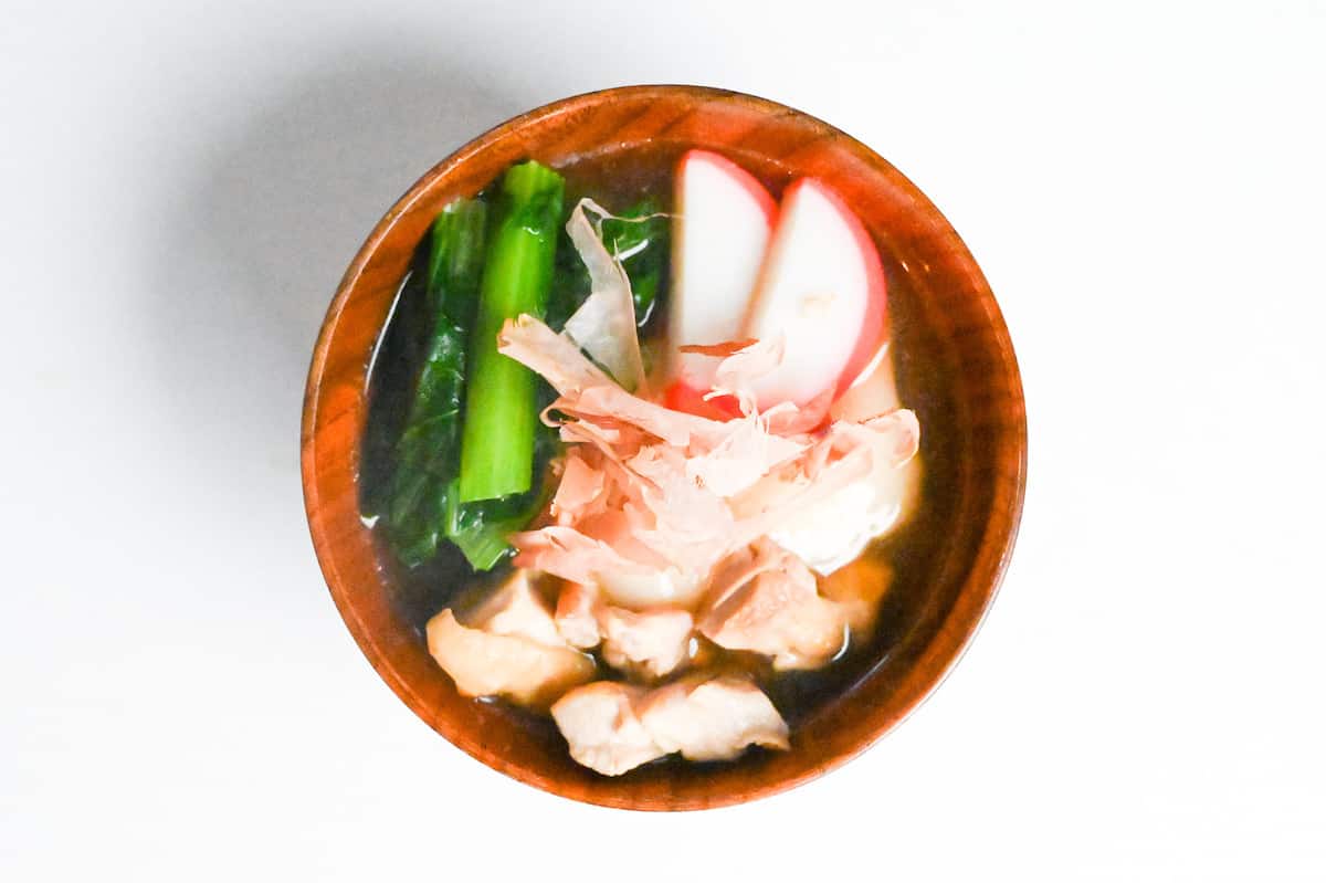 ozoni in a wooden bowl topped with pink and white fishcakes (kamaboko), blanched komatsuna and bonito flakes