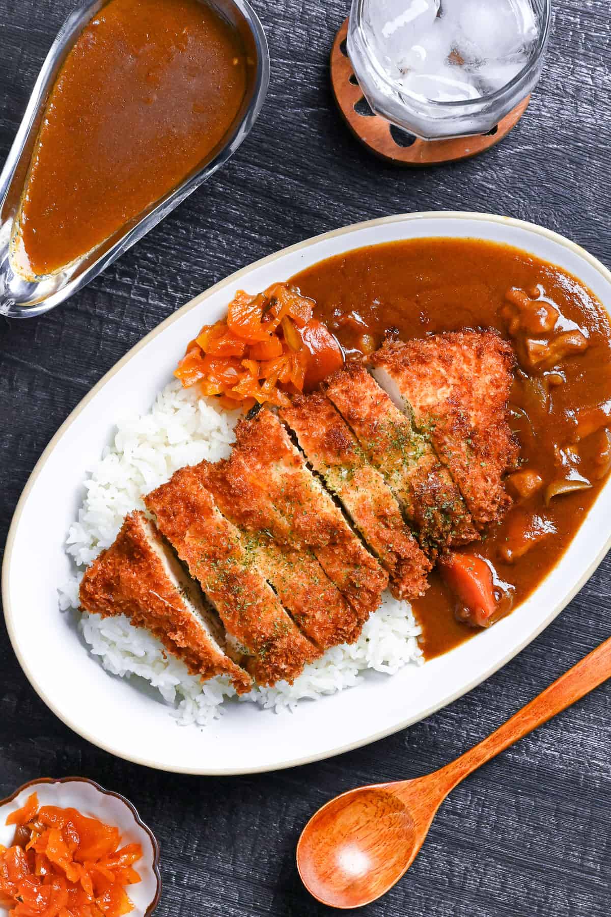 Japanese chicken katsu curry served on a white oval-shaped plate with beige rim, next to a wooden spoon, bowl of pickles and gravy boat of curry on a black background