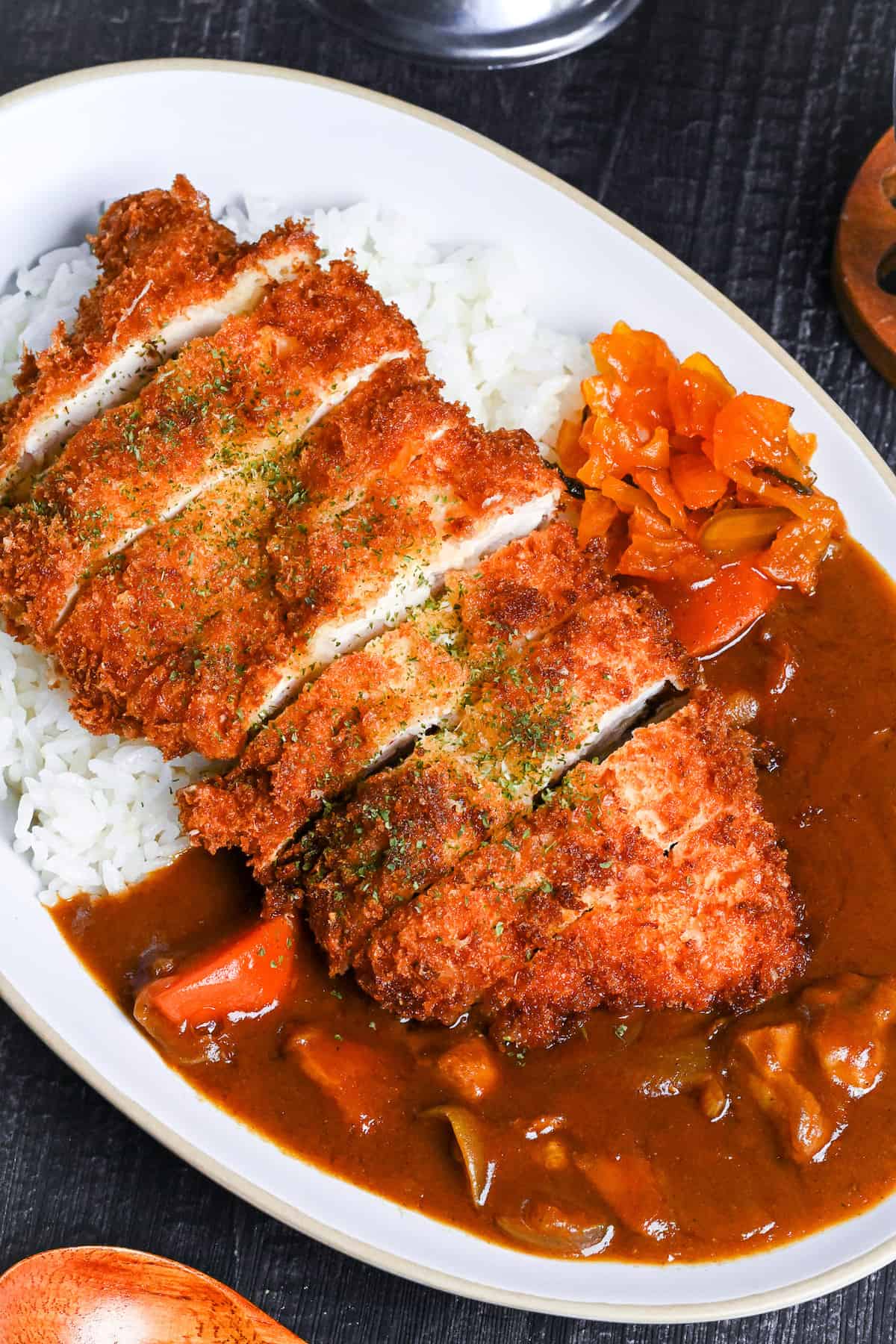 Japanese chicken katsu curry served on a white oval-shaped plate with beige rim, next to a wooden spoon, bowl of pickles and gravy boat of curry on a black background