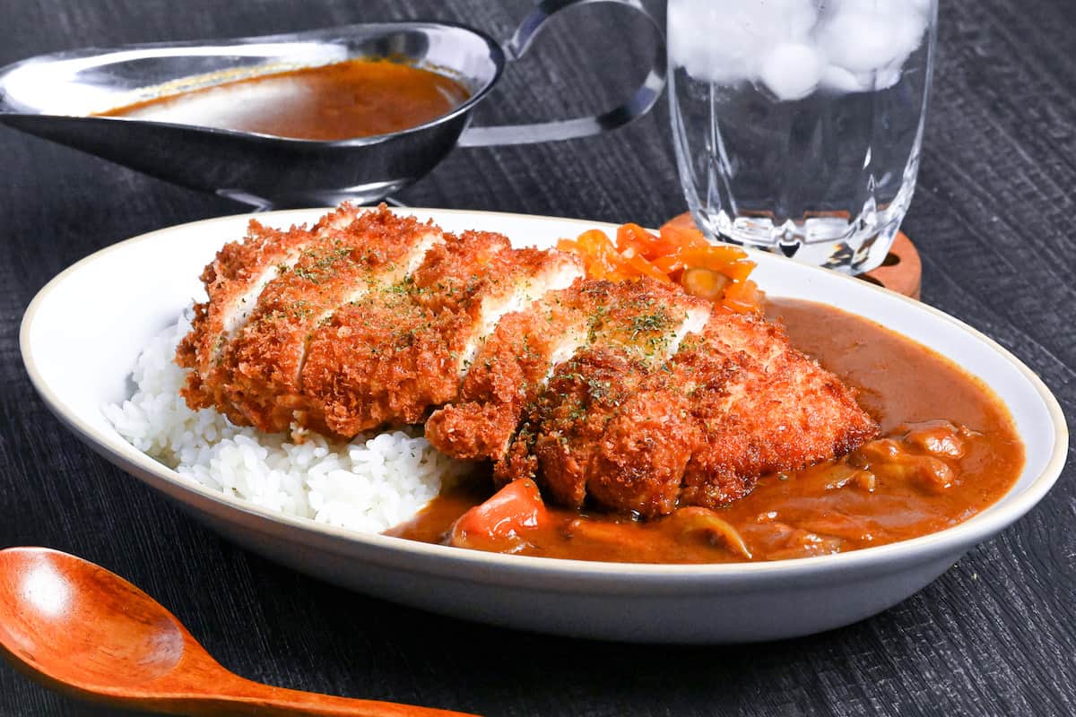 Japanese chicken katsu curry served on a white oval-shaped plate with beige rim, next to a wooden spoon, glass of water and gravy boat of curry on a black background