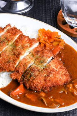 Japanese chicken katsu curry served on a white oval-shaped plate with beige rim, next to a wooden spoon on a black background topdown view