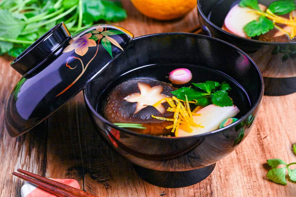 Japanese Clear Soup (Osuimono) in a black bowl with decorative lid, made with shiitake mushroom, kamaboko fish cake, dried wheat gluten (temari fu), yuzu peel and Japanese wild parsley