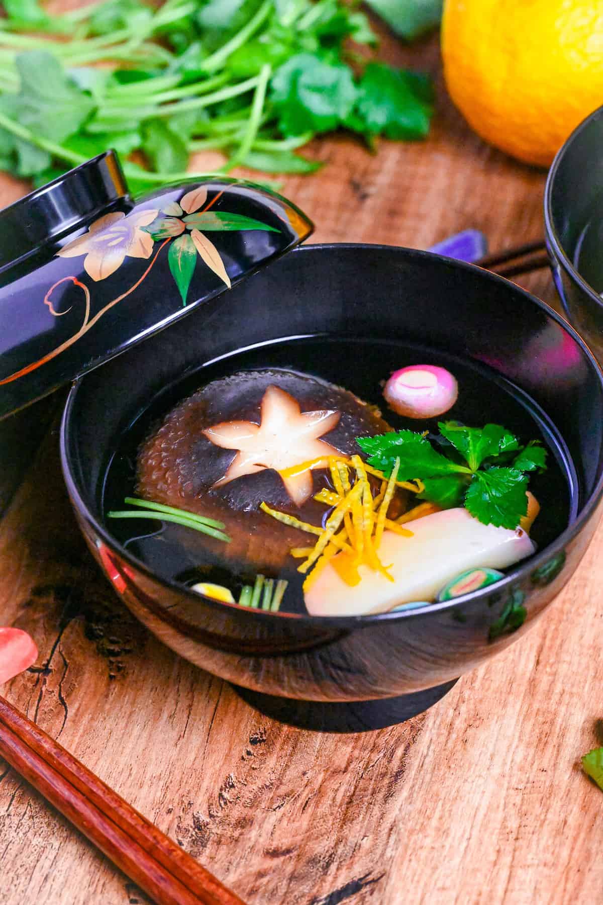 Japanese Clear Soup (Osuimono) in a black bowl with decorative lid, made with shiitake mushroom, kamaboko fish cake, dried wheat gluten (temari fu), yuzu peel and Japanese wild parsley