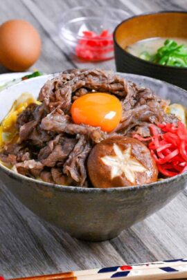 Sukiyaki beef donburi rice bowl topped with red pickled ginger, shiitake mushroom and egg yolk in a mottled bowl