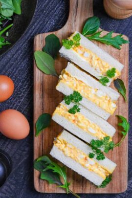 Four Japanese egg finger sandwiches (tamago sando) on a wooden chopping board with salad leaves