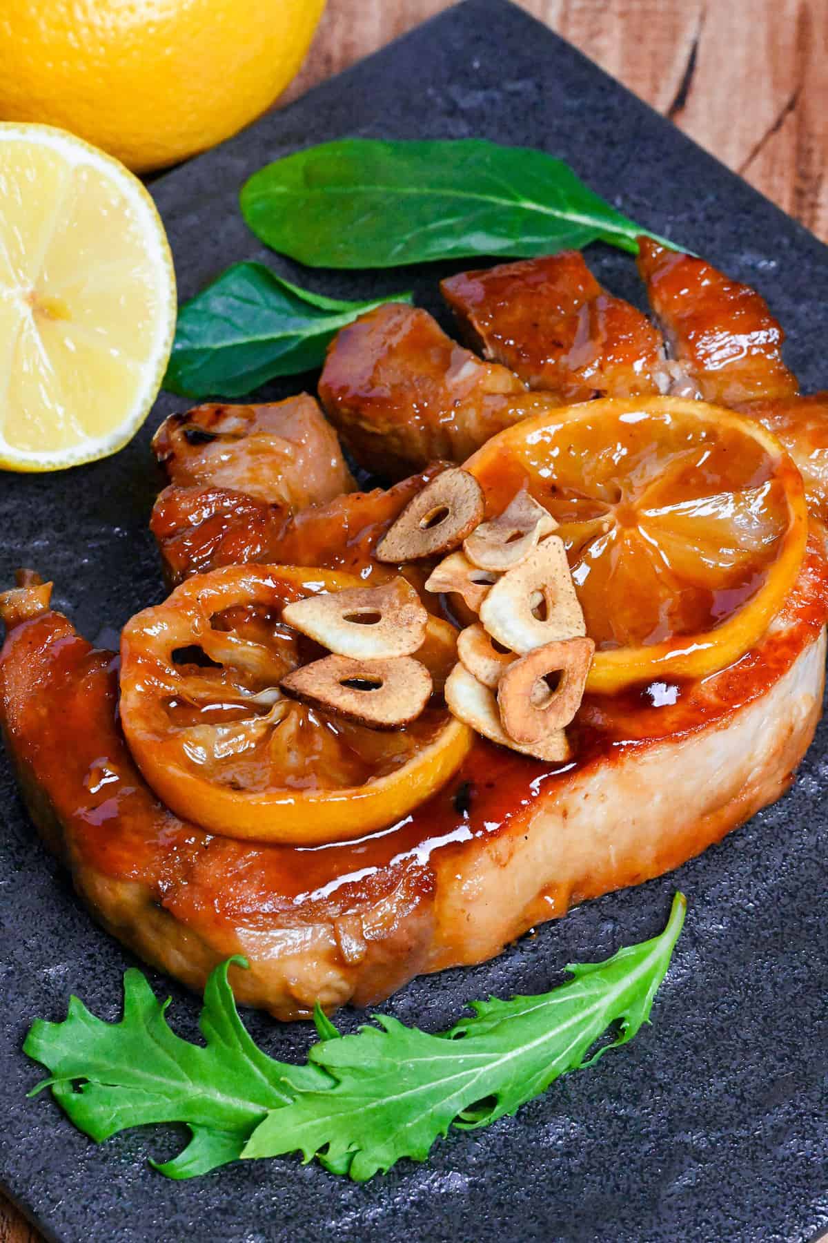 Lemon Teriyaki Pork Chop topped with lemon slices and fried garlic chips on a black slate-style square plate with pan fried mushrooms and salad leaves