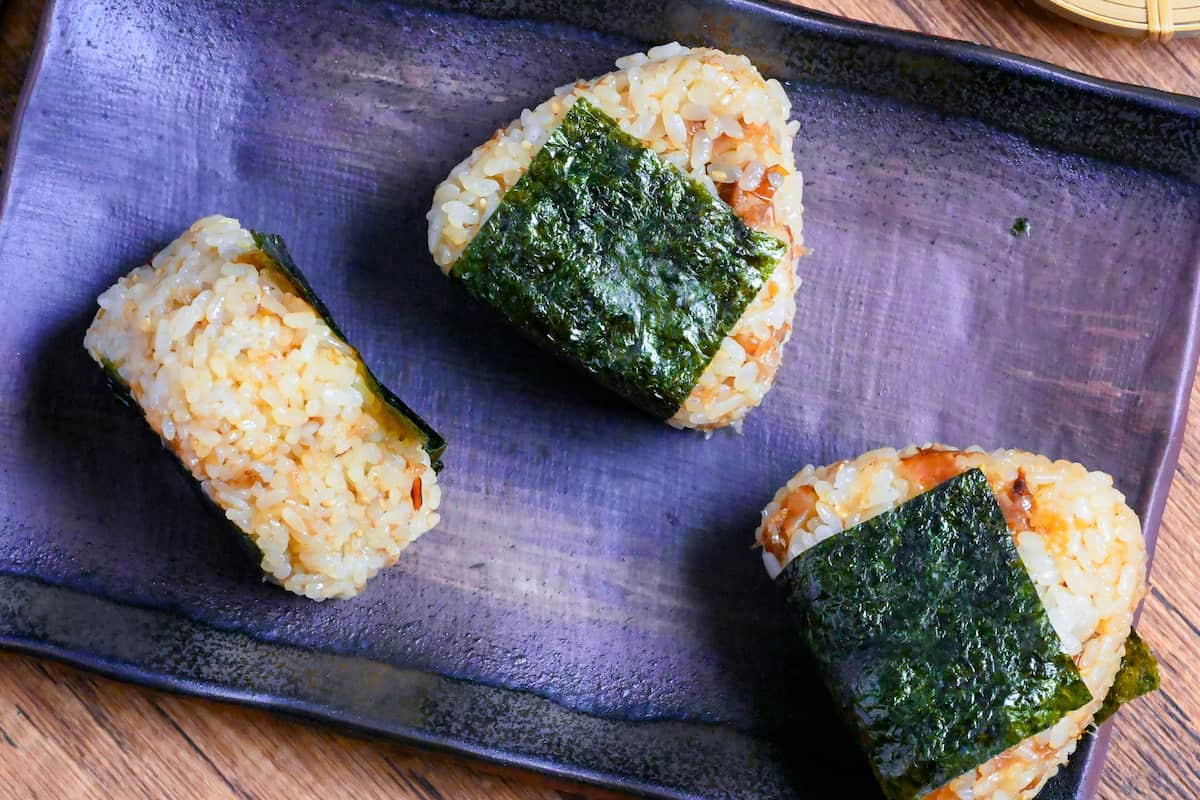 3 okaka onigiri (seasoned bonito flake rice balls) lined up on a dark brown rectangular plate on a wooden background