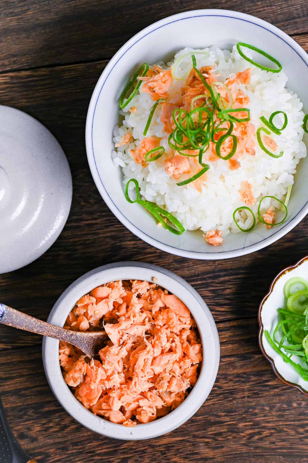 Homemade Japanese-style salmon flakes in a white ceramic pot with brown decorative spoon next to a bowl of rice and small dish of green onions