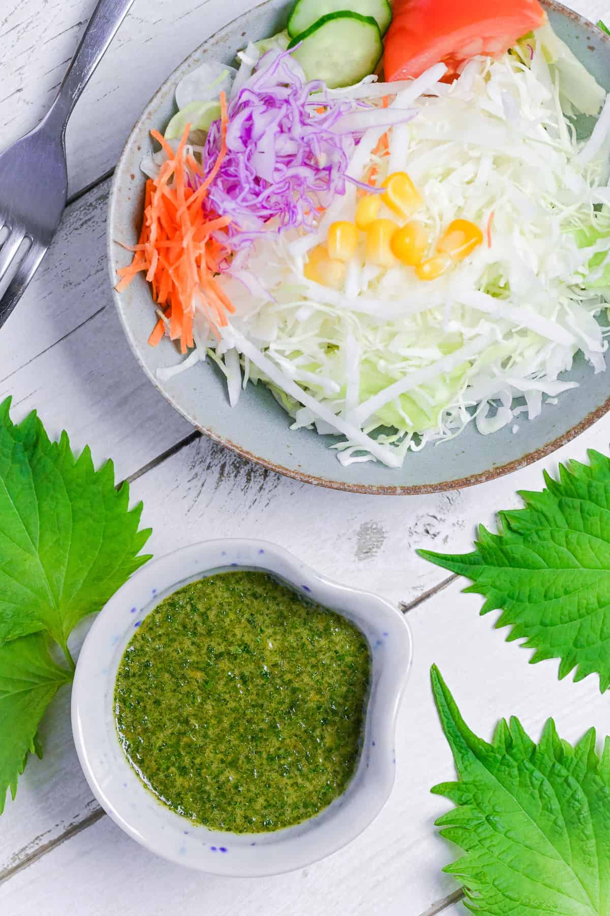 Japanese shiso dressing in a small blue and brown ceramic jug with a bowl of salad in the background and shiso leaves scattered around