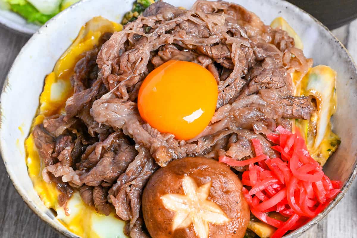 Sukiyaki beef donburi rice bowl topped with red pickled ginger, shiitake mushroom and egg yolk in a mottled bowl close up