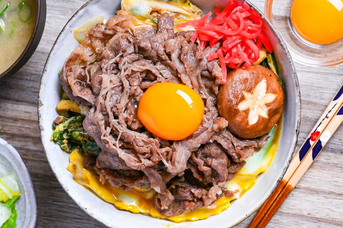Sukiyaki beef donburi rice bowl topped with red pickled ginger, shiitake mushroom and egg yolk in a mottled bowl top down close up