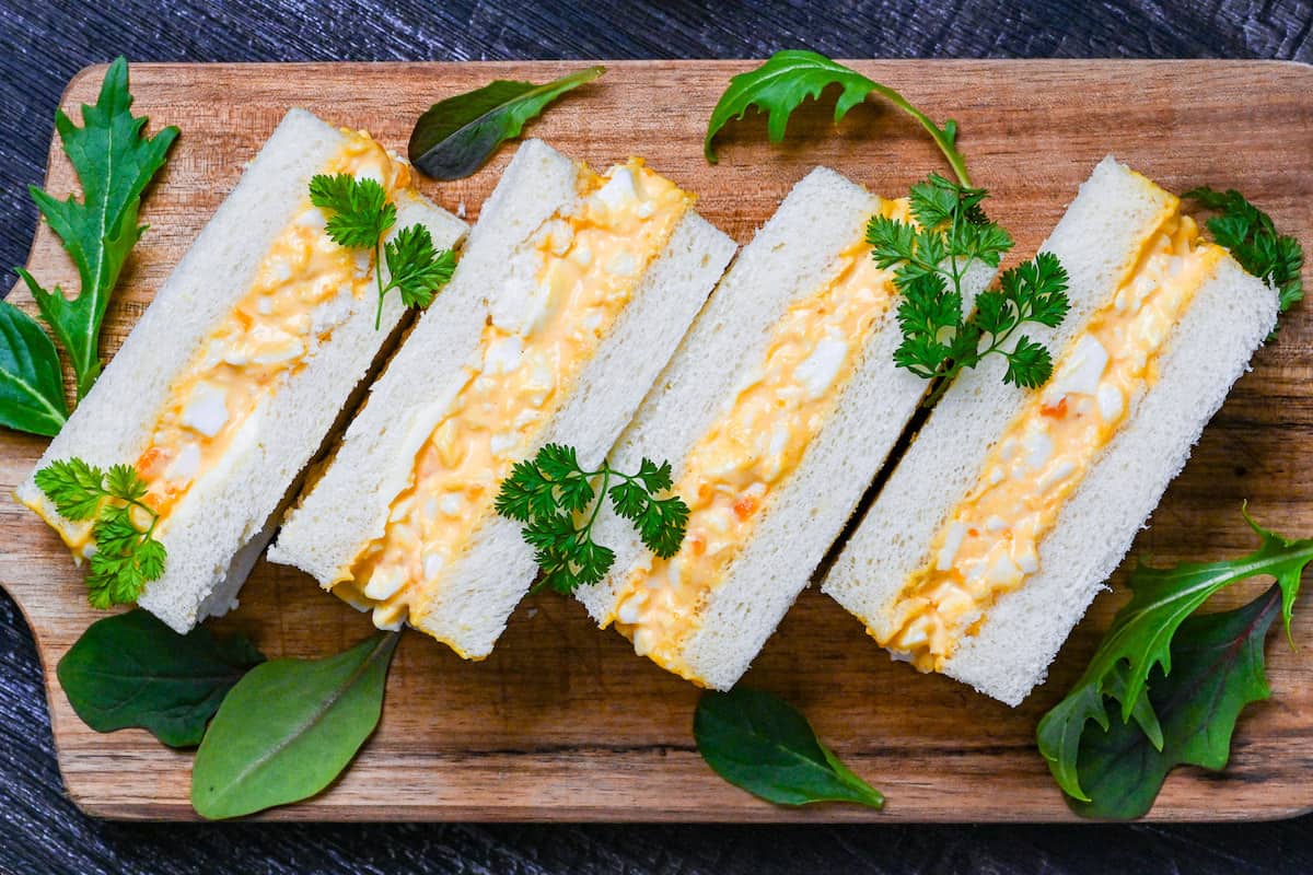 Four Japanese egg finger sandwiches (tamago sando) on a wooden chopping board with salad leaves