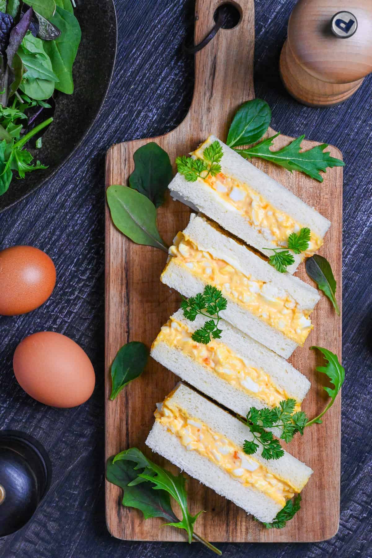 Four Japanese egg finger sandwiches (tamago sando) on a wooden chopping board with salad leaves