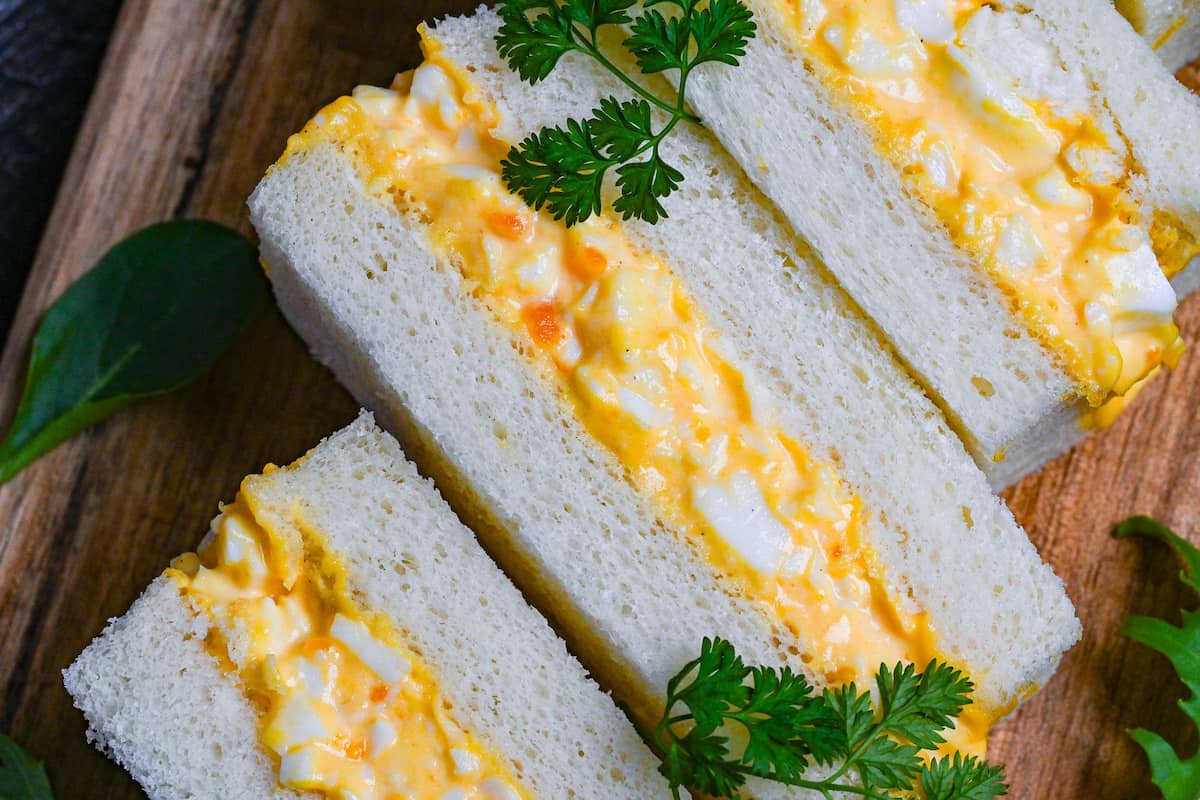Four Japanese egg finger sandwiches (tamago sando) on a wooden chopping board with salad leaves