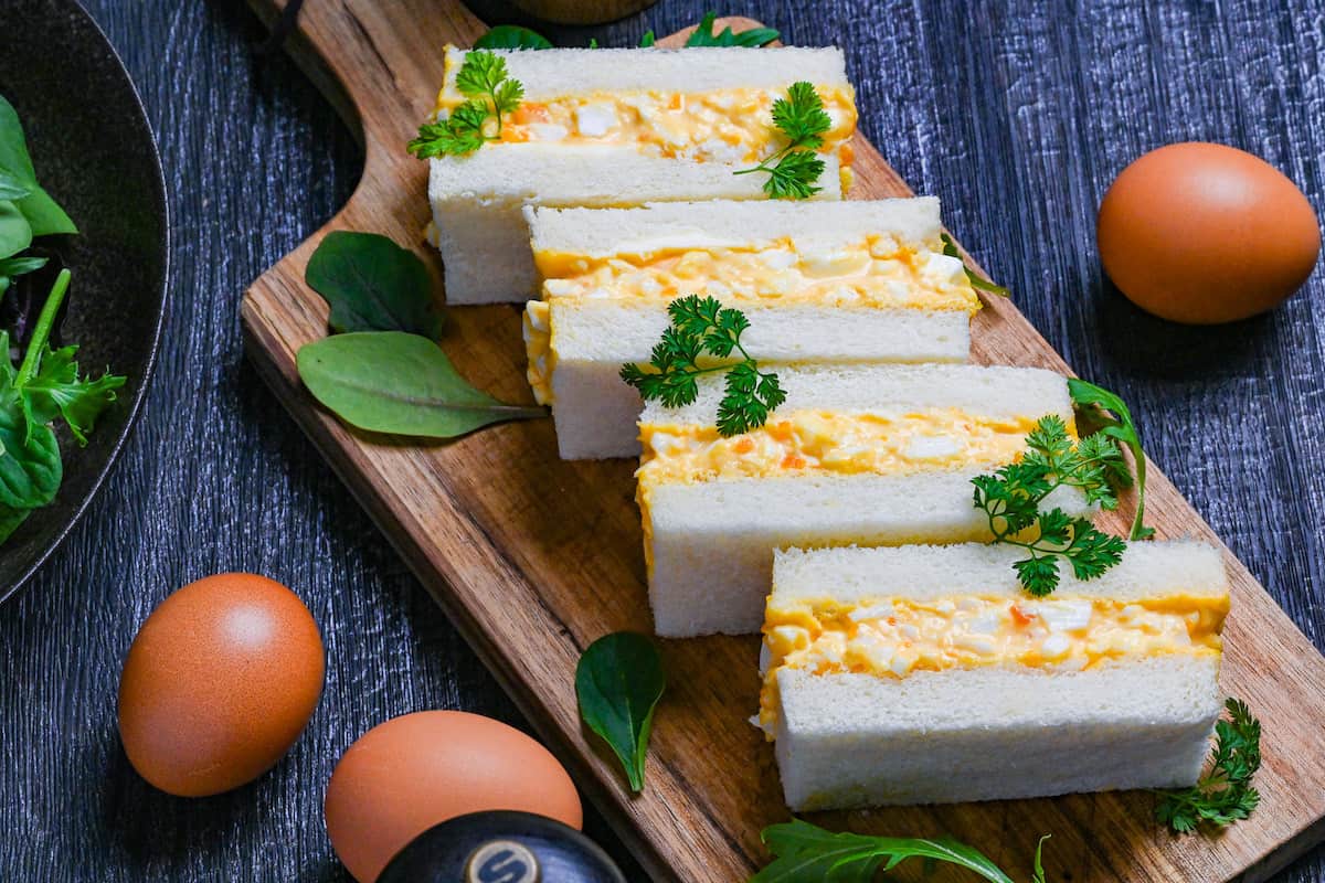 Four Japanese egg finger sandwiches (tamago sando) on a wooden chopping board with salad leaves