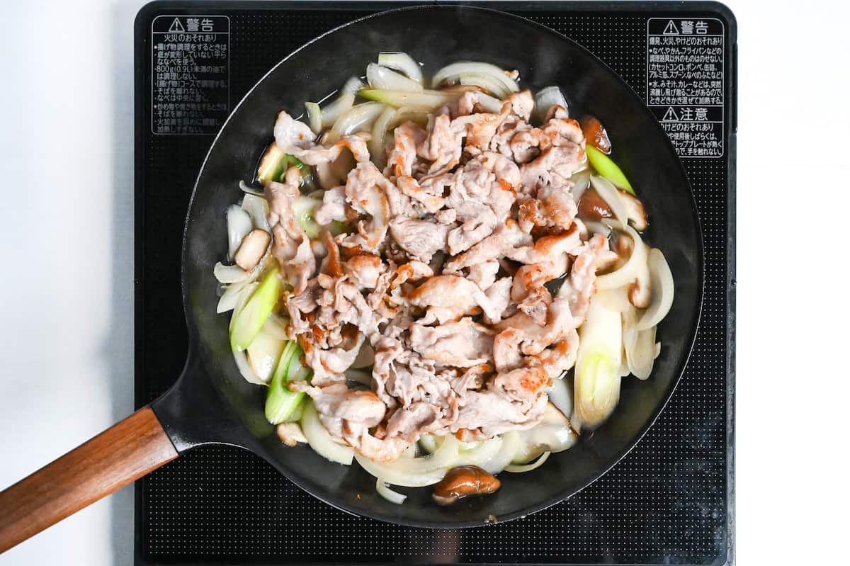 cooked thinly sliced pork with Japanese leek, yellow onions and shiitake mushroom slices in a black frying pan