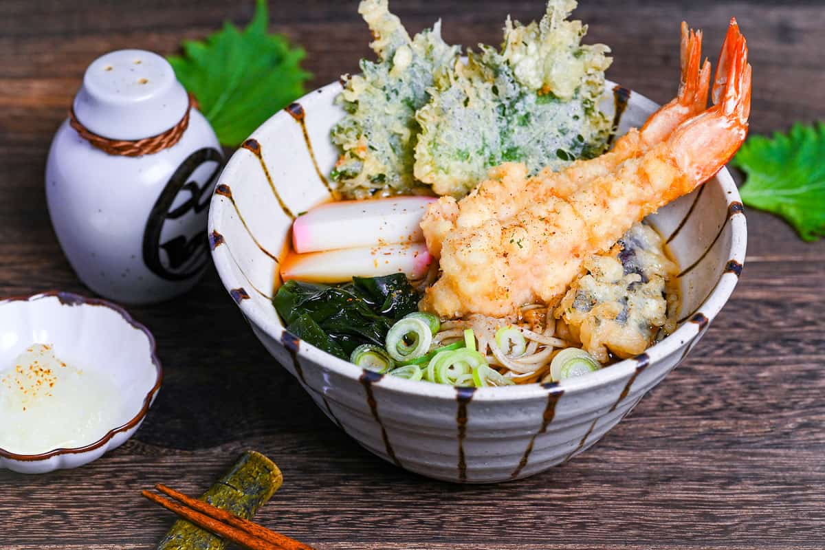 Tempura soba topped with tempura shrimp and vegetables, chopped green onions, kamaboko fishcakes and wakame seadweed in a striped bowl on a dark wooden effect background
