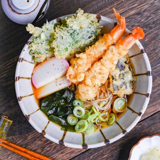 Tempura soba topped with tempura shrimp and vegetables, chopped green onions, kamaboko fishcakes and wakame seadweed in a striped bowl on a dark wooden effect background top down view