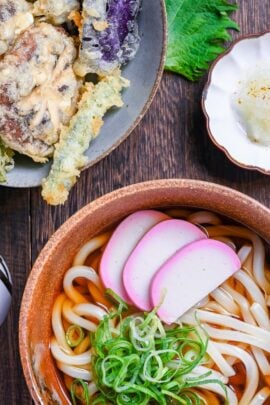 Udon noodles in simple broth in a brown bowl next to a variety of vegetable tempura including shiitake mushrooms, asparagus, shiso leaves and eggplant