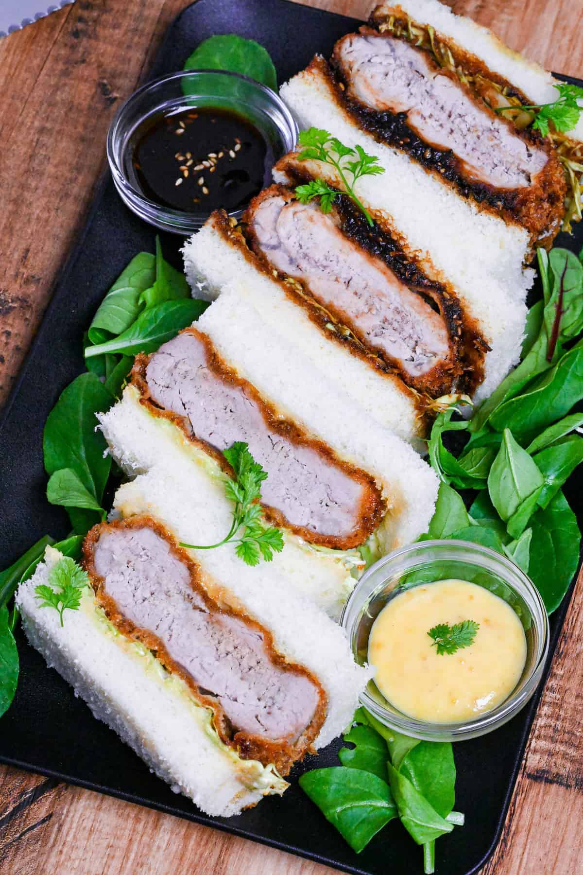 4 katsu sando made with two kinds of sauce on a black plate surrounded by baby leaf salad on a wood-effect background