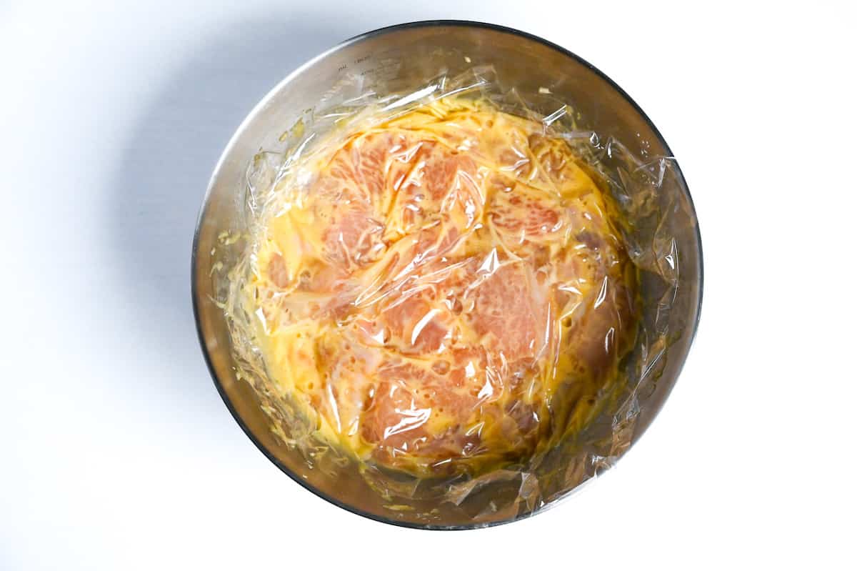 chicken breast marinating in egg and condiments in a mixing bowl covered with plastic wrap