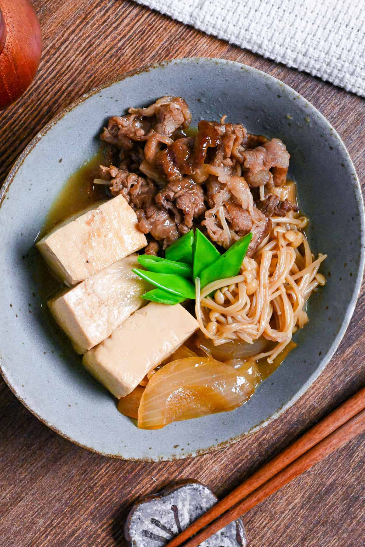 niku dofu in a eggshell blue bowl made with firm tofu, thinly sliced beef, onions and enoki mushrooms simmered in a sweet and salty Japanese broth and topped with blanched snow peas on a wooden table