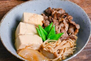 niku dofu in a eggshell blue bowl made with firm tofu, thinly sliced beef, onions and enoki mushrooms simmered in a sweet and salty Japanese broth and topped with blanched snow peas on a wooden table
