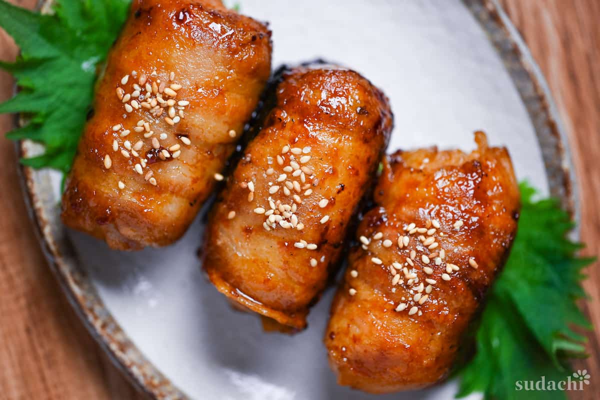 Three nikumaki onigiri (pork belly wrapped rice balls) arranged on a cream plate decorated with Perilla leaves and sprinkled with sesame seeds