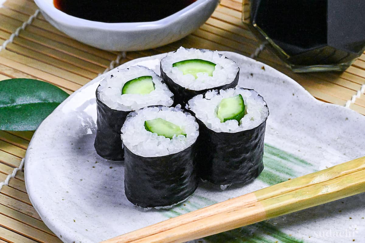 4 kappa maki (cucumber sushi rolls) on a gray plate with green brush effect on a bamboo sushi mat next to a white bowl of soy sauce