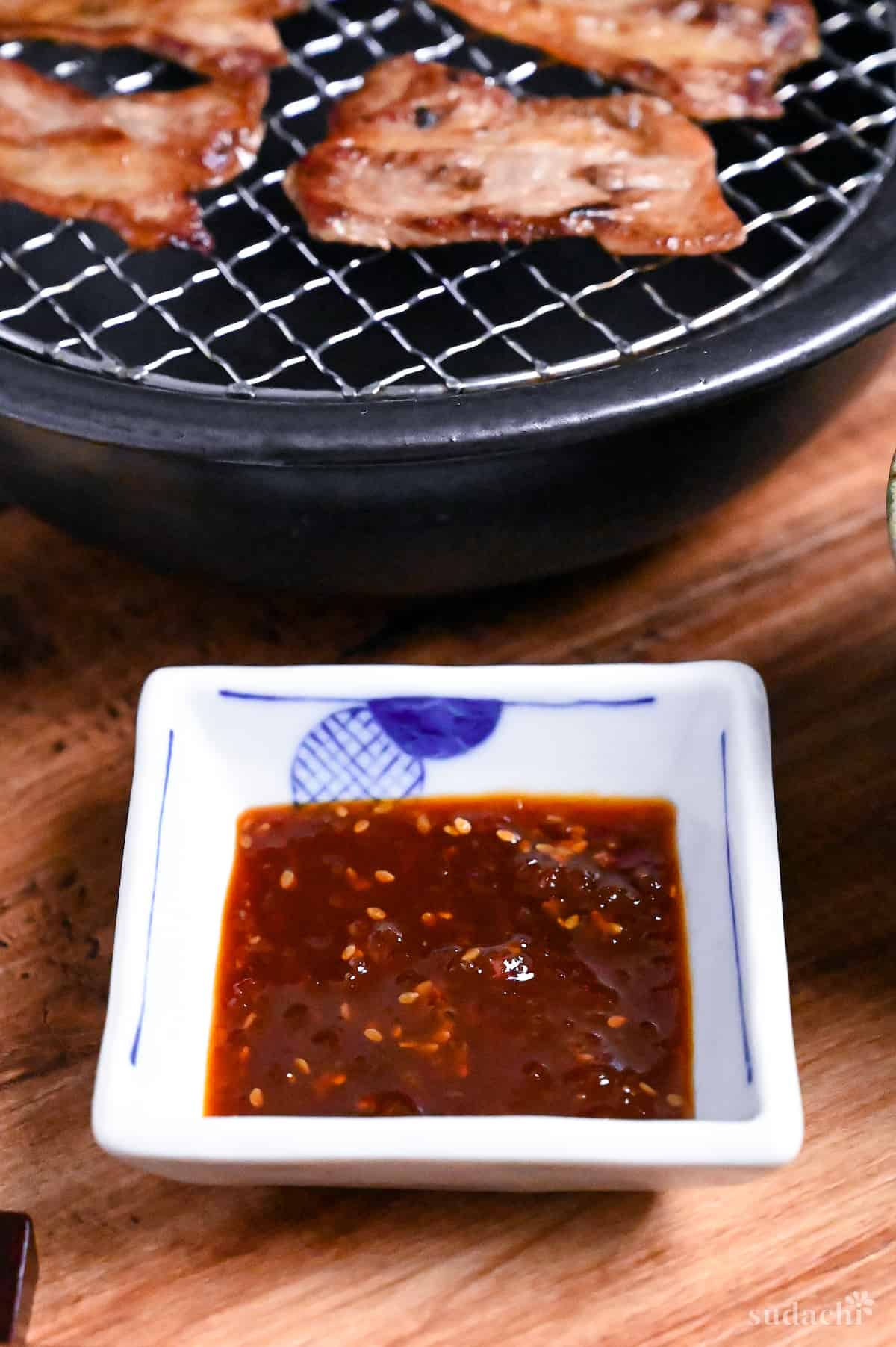 Homemade Japanese yakiniku sauce in a small square white bowl with blue border next to slices of thinly sliced beef on a metal grill