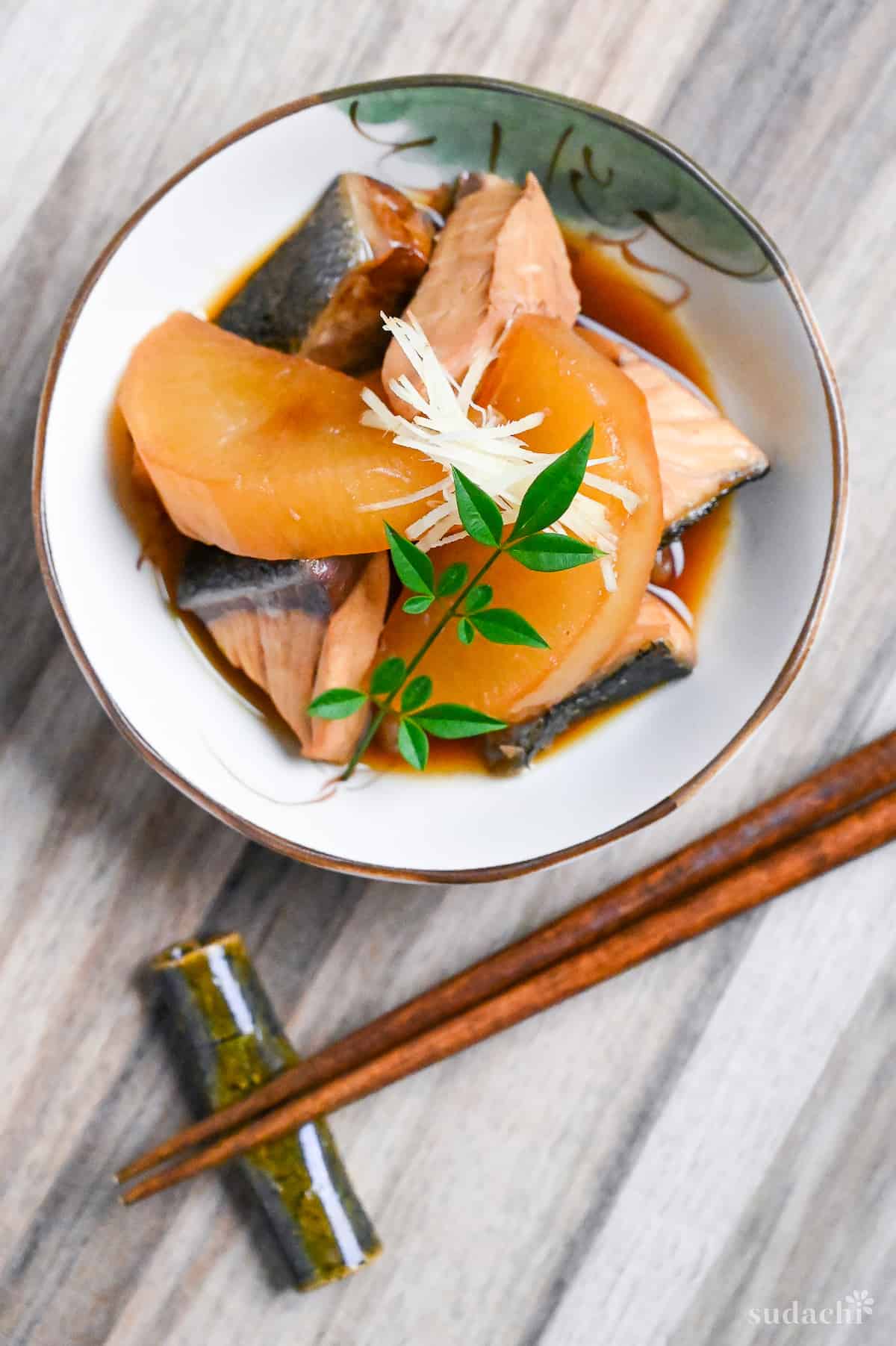 Buri Daikon (Simmered Yellowtail and Daikon Radish) in a small cream bowl with Japanese design topped with decorative leaves