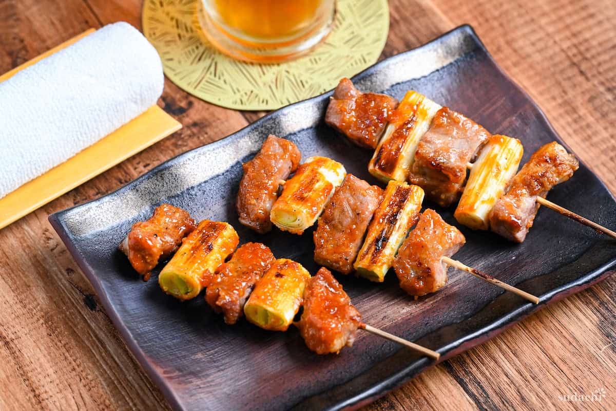 Three Yakiton skewers made with grilled pork shoulder and Japanese leek served on a skewer and brushed with a miso tare (sauce) on a brown rectangular plate