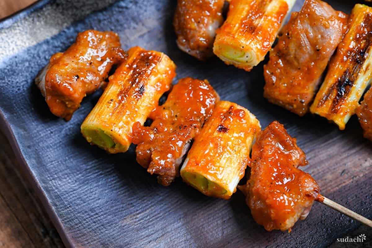 Three Yakiton skewers made with grilled pork shoulder and Japanese leek served on a skewer and brushed with a miso tare (sauce) on a brown rectangular plate
