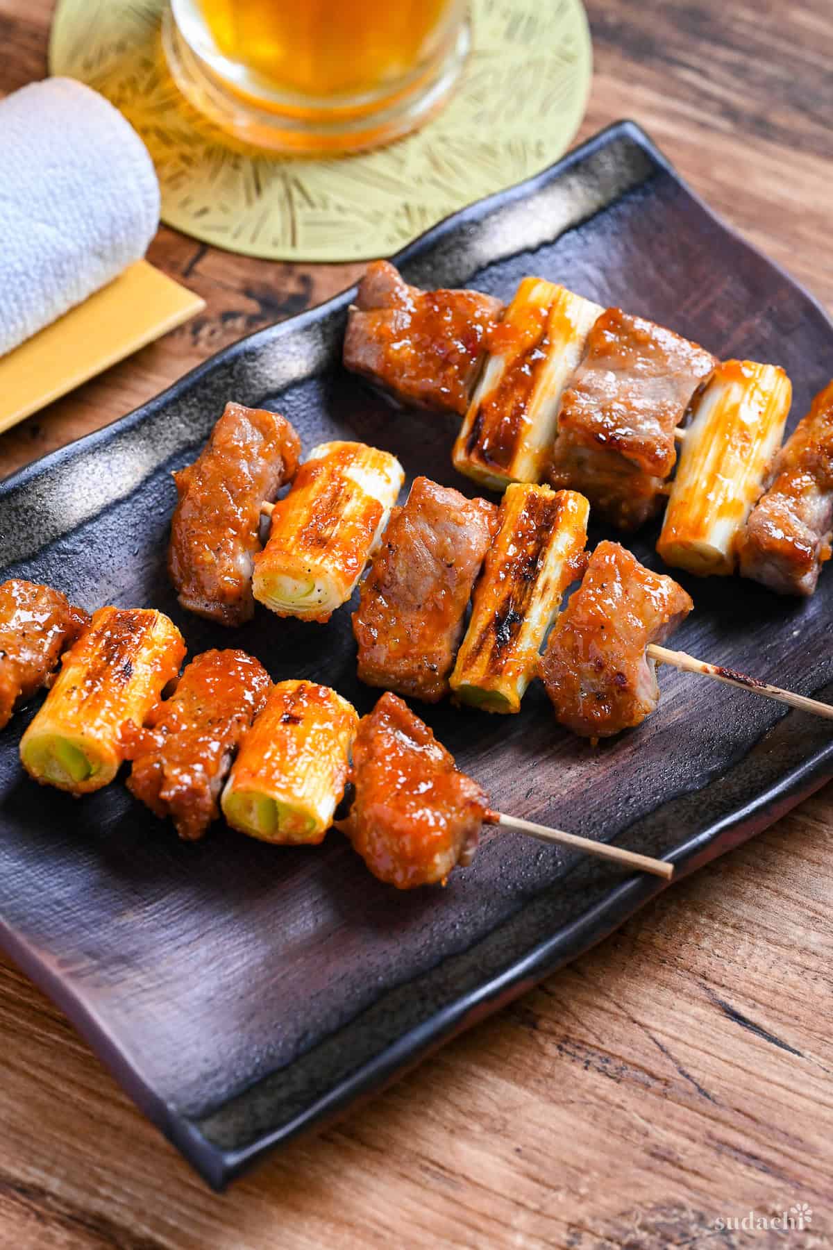 Three Yakiton skewers made with grilled pork shoulder and Japanese leek served on a skewer and brushed with a miso tare (sauce) on a brown rectangular plate