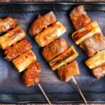 Three Yakiton skewers made with grilled pork shoulder and Japanese leek served on a skewer and brushed with a miso tare (sauce) on a brown rectangular plate