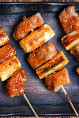 Three Yakiton skewers made with grilled pork shoulder and Japanese leek served on a skewer and brushed with a miso tare (sauce) on a brown rectangular plate