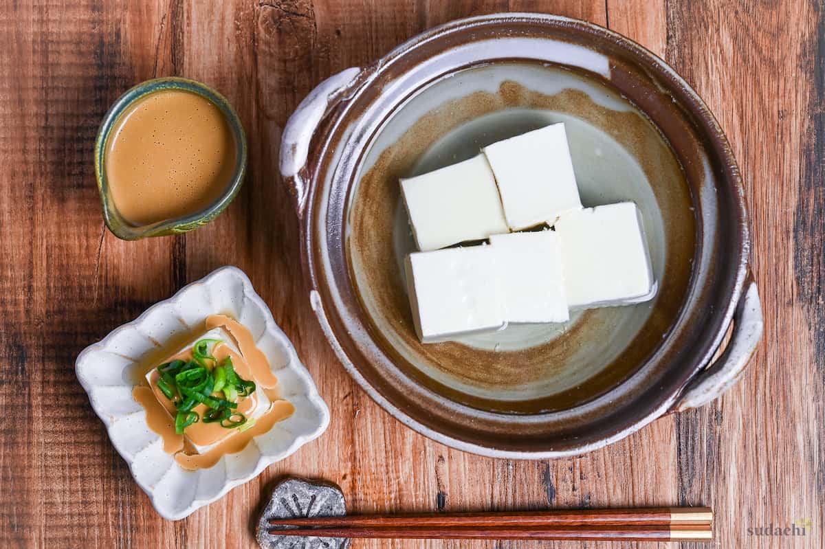Yudofu simmered tofu drizzled with homemade sesame sauce topped with chopped green onions in a square white fluted bowl next to a pot of simmered tofu and a small jug of sauce