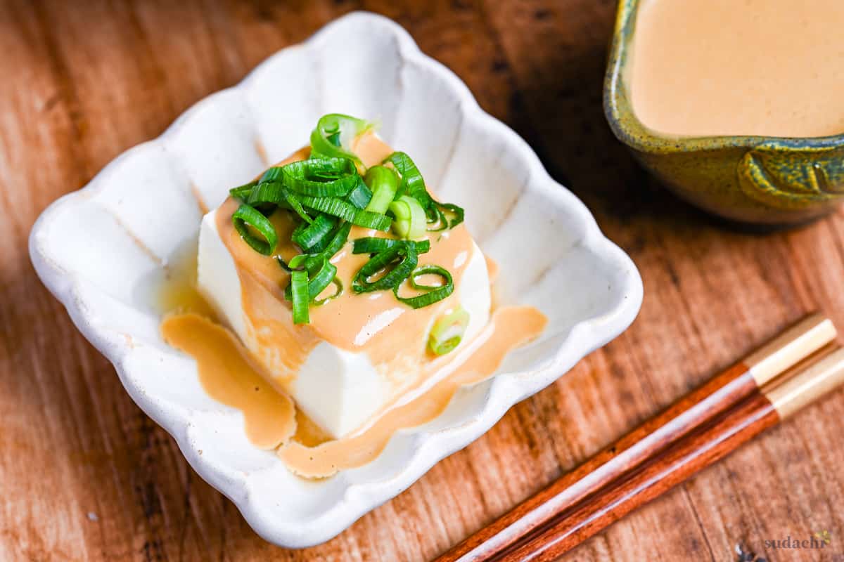 Yudofu simmered tofu drizzled with homemade sesame sauce topped with chopped green onions in a square white fluted bowl