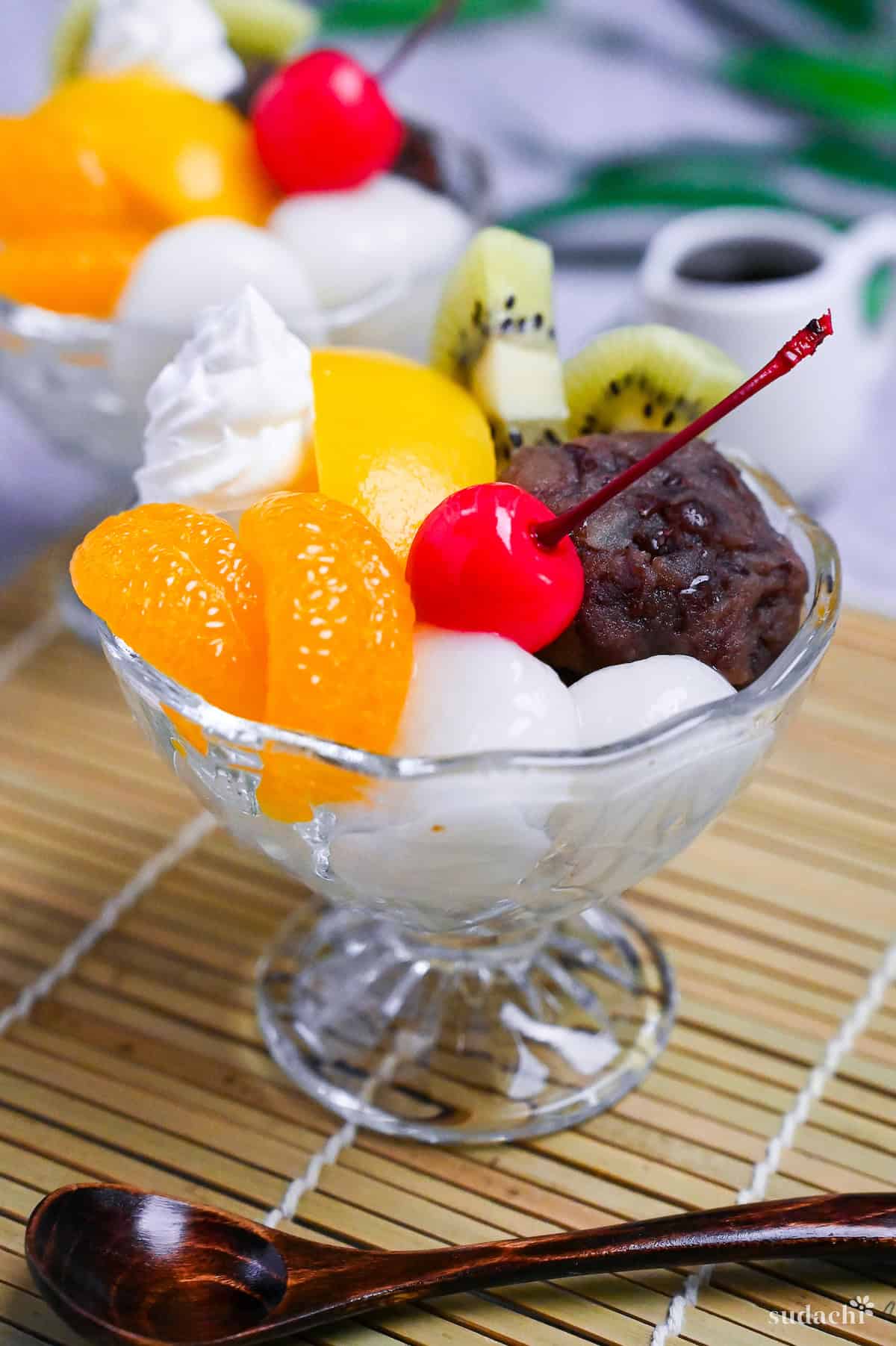 Anmitsu (Japanese kanten jelly dessert with red bean paste) topped with mikan orange, peach, kiwi, shiratama dangos, a cherry and whipped cream next to a small jug of homemade kuromitsu syrup