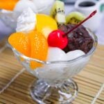 Anmitsu (Japanese kanten jelly dessert with red bean paste) topped with mikan orange, peach, kiwi, shiratama dangos, a cherry and whipped cream next to a small jug of homemade kuromitsu syrup