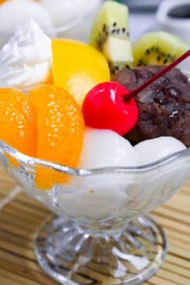 Anmitsu (Japanese kanten jelly dessert with red bean paste) topped with mikan orange, peach, kiwi, shiratama dangos, a cherry and whipped cream next to a small jug of homemade kuromitsu syrup