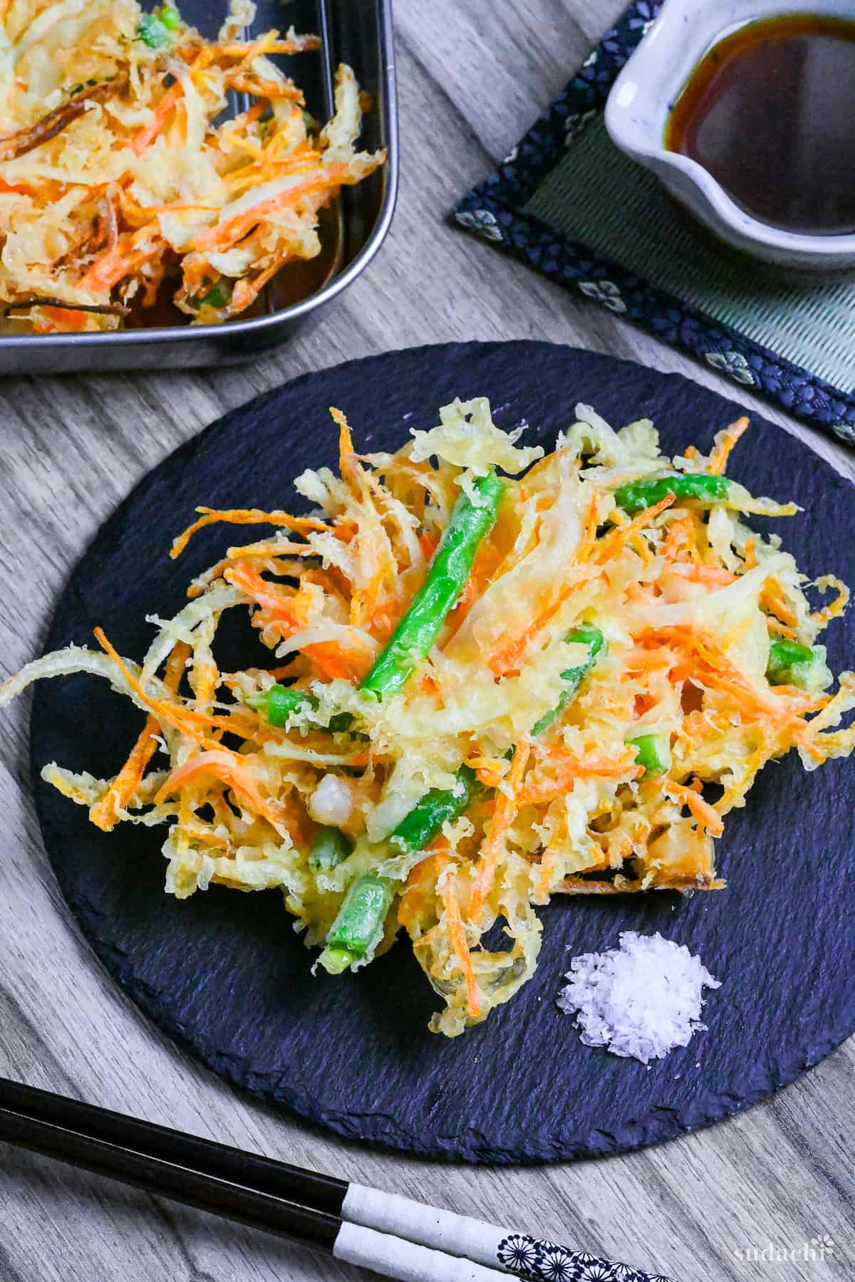 Kakiage (Japanese mixed vegetable tempura) on a slate plate next to a small heap of coarse salt for dipping and a jug of dipping sauce in the background