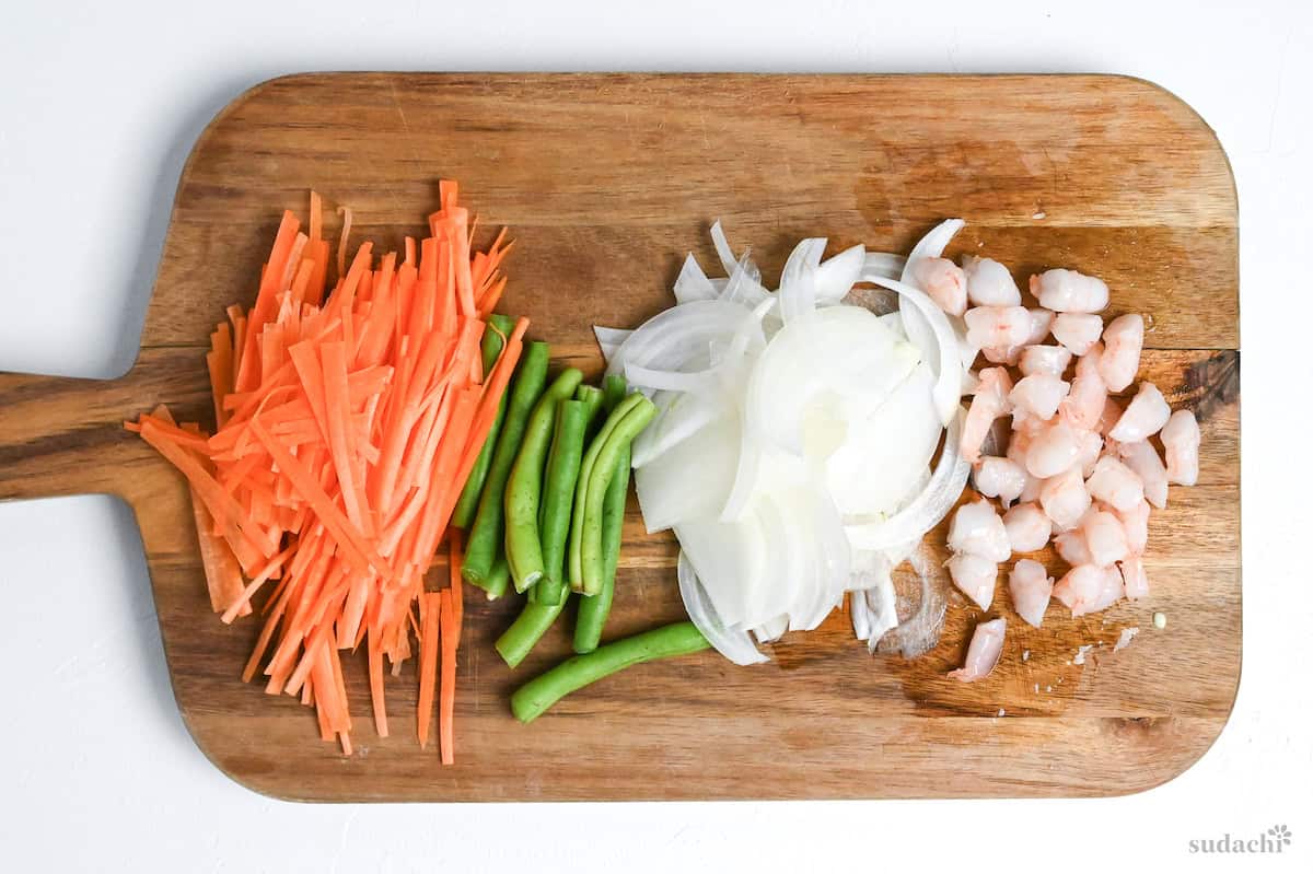 julienned carrot, sliced runner beans, thinly sliced onion and roughly chopped boiled shrimp on a wooden chopping board on a white background