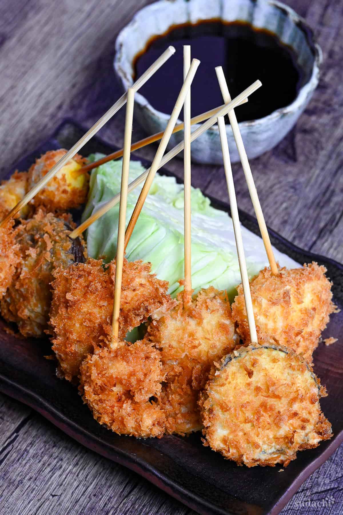 Kushi age/Kushi katsu battered skewers on a brown rectangular plate resting on a wedge of cabbage with dipping sauce in the background