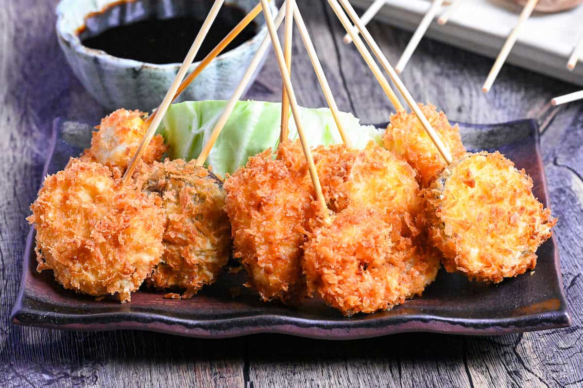 Kushi age/Kushi katsu battered skewers on a brown rectangular plate resting on a wedge of cabbage with dipping sauce in the background