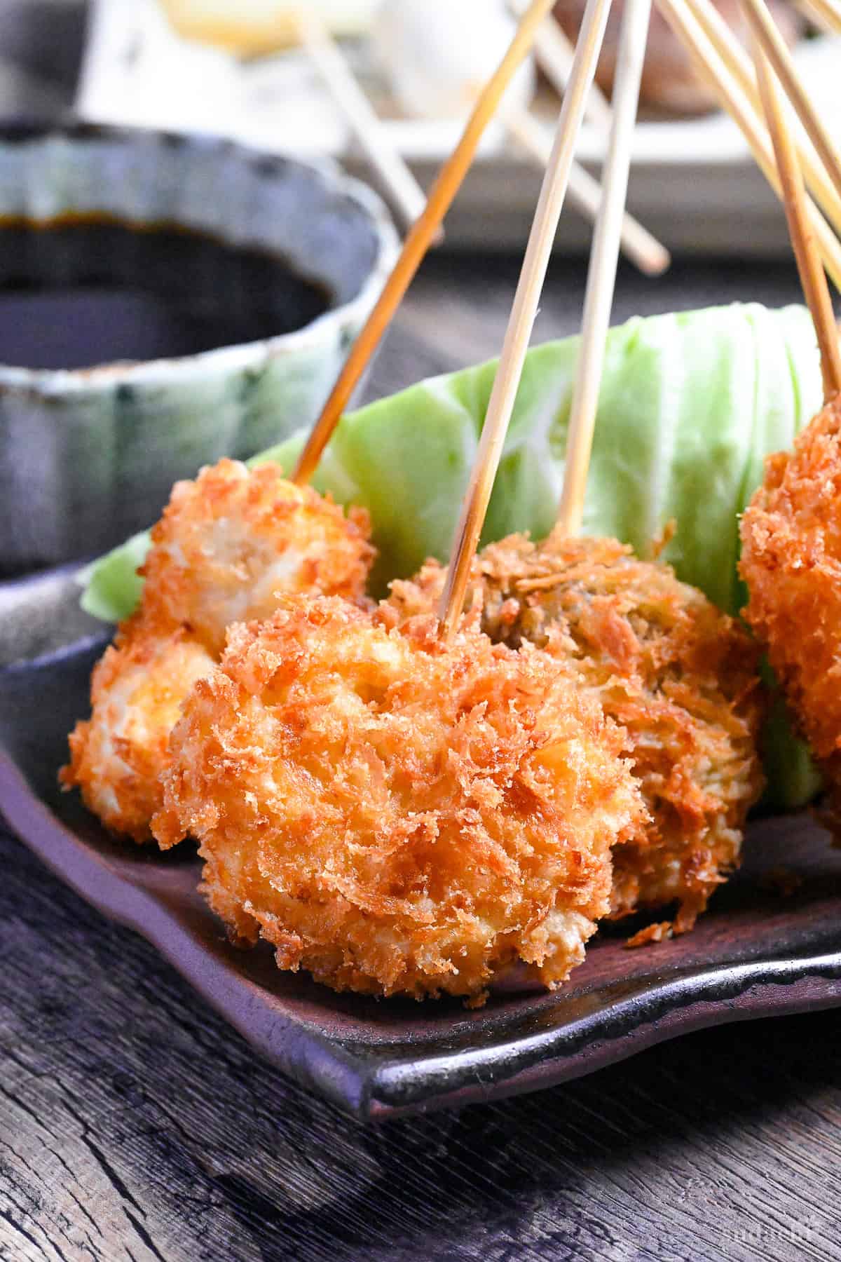 Kushi age/Kushi katsu battered skewers on a brown rectangular plate resting on a wedge of cabbage with dipping sauce in the background