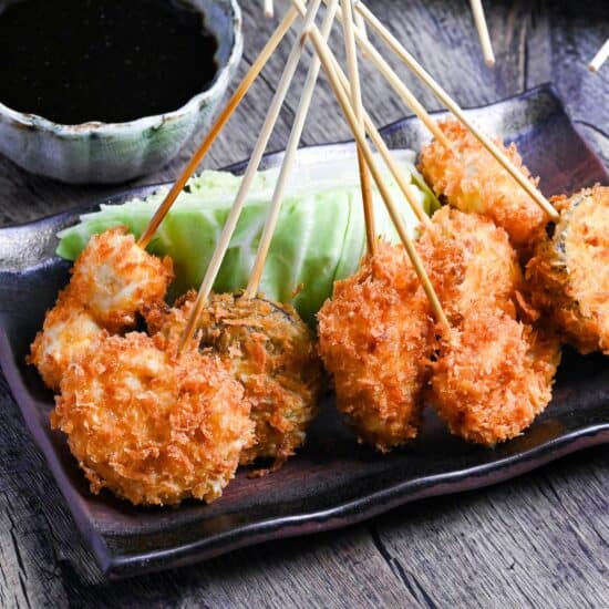 Kushi age/Kushi katsu battered skewers on a brown rectangular plate resting on a wedge of cabbage with dipping sauce in the background