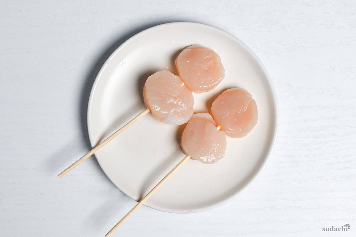 4 raw scallops on bamboo skewers on a white plate on a white background